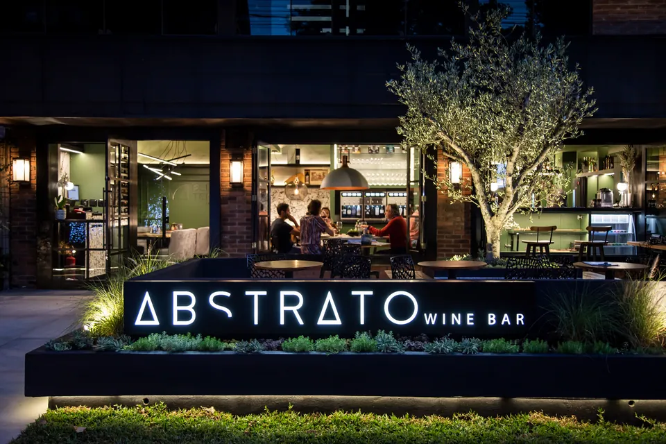 Fachada externa noturna do Abstrato Wine Bar em Gramado com letreiro iluminado, deck com oliveira e clientes no salão acolhedor.
